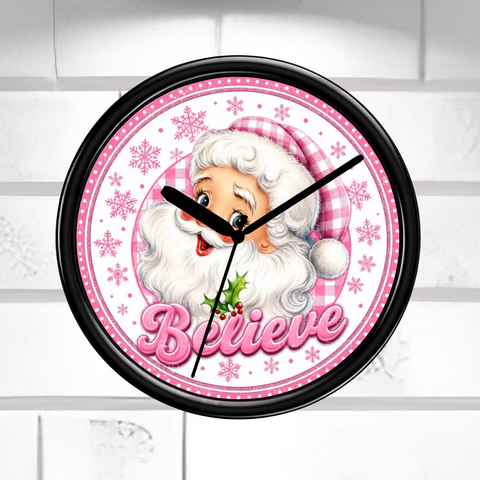 A round wall clock with a black frame and a festive Santa Claus face on the dial. The clock face features pink gingham patterns, pink snowflakes, the word 'Believe' in pink script, and black clock hands against a light gray tiled wall background.