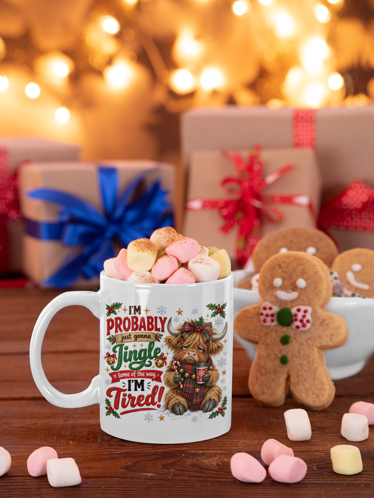 A white ceramic mug filled with marshmallows sits on a wooden table in a festive holiday setting. The mug features a humorous Christmas graphic with a highland cow and the text 'I'M PROBABLY just going 'Jingle Wisdom of the year I'm Tired!'.