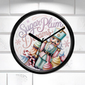 A round wall clock with a black frame and a whimsical nutcracker illustration on the face. The clock face features the text 'Sugar Plum Dreams' in purple script, along with cupcakes, candy canes, snowflakes, and pastel-colored details.