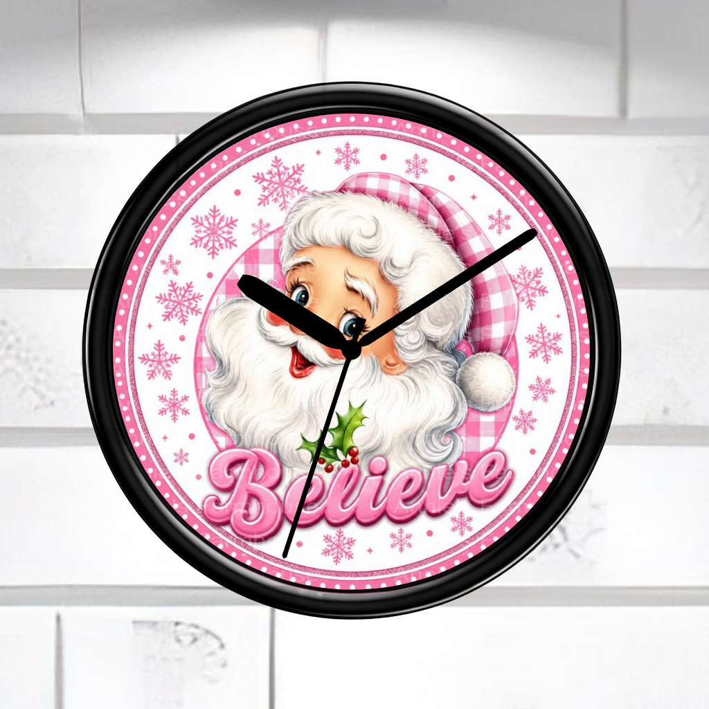 A round wall clock with a black frame and a festive Santa Claus face on the dial. The clock face features pink gingham patterns, pink snowflakes, the word 'Believe' in pink script, and black clock hands against a light gray tiled wall background.