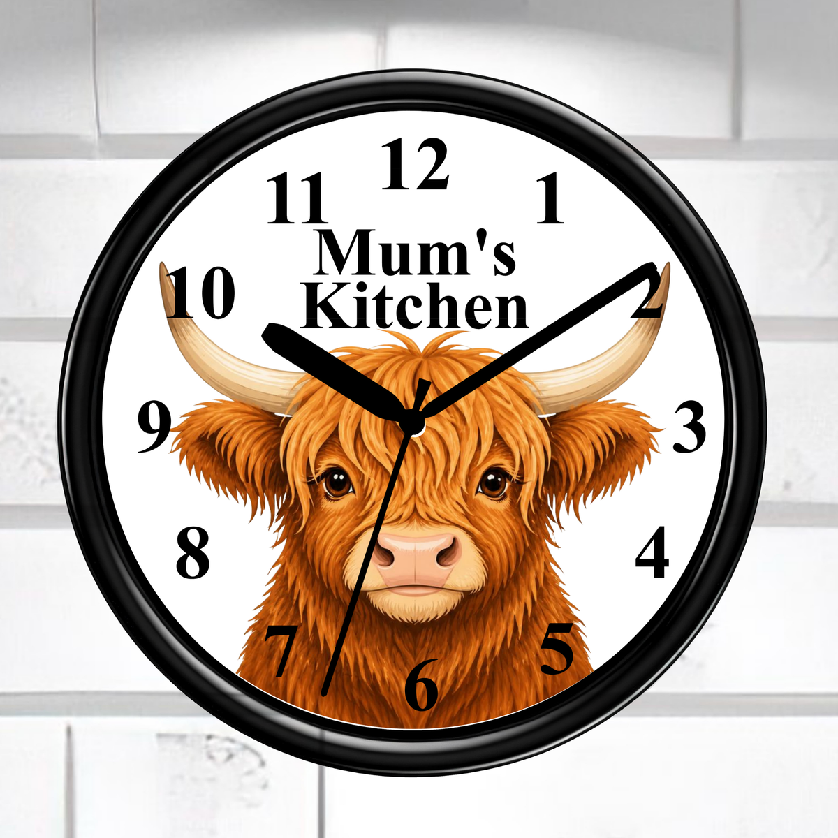 A round wall clock with a black frame and a white face, featuring a brown highland cow illustration and the text 'Mum's Kitchen' at the center. The clock is mounted against a white tiled wall.