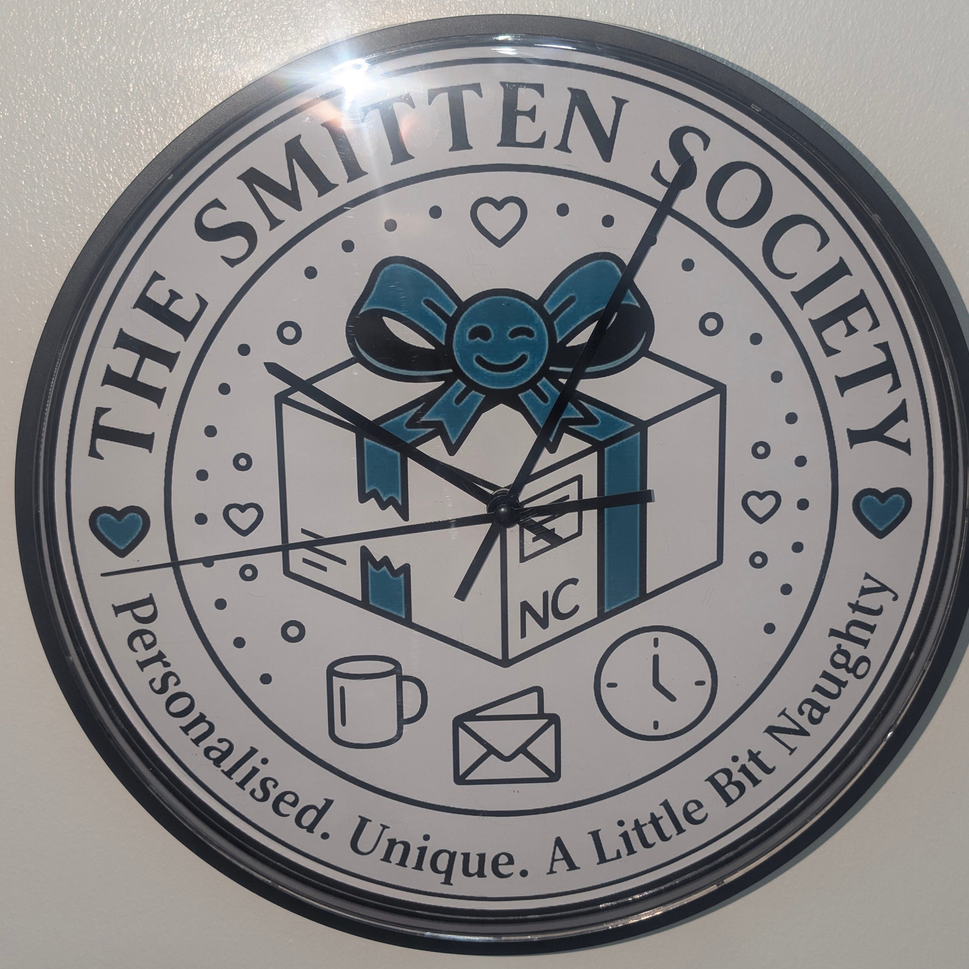 A round novelty wall clock featuring a white face with a black outer rim and printed design. The design includes the text 'THE SMITTEN SOCIETY' and 'Personalised.