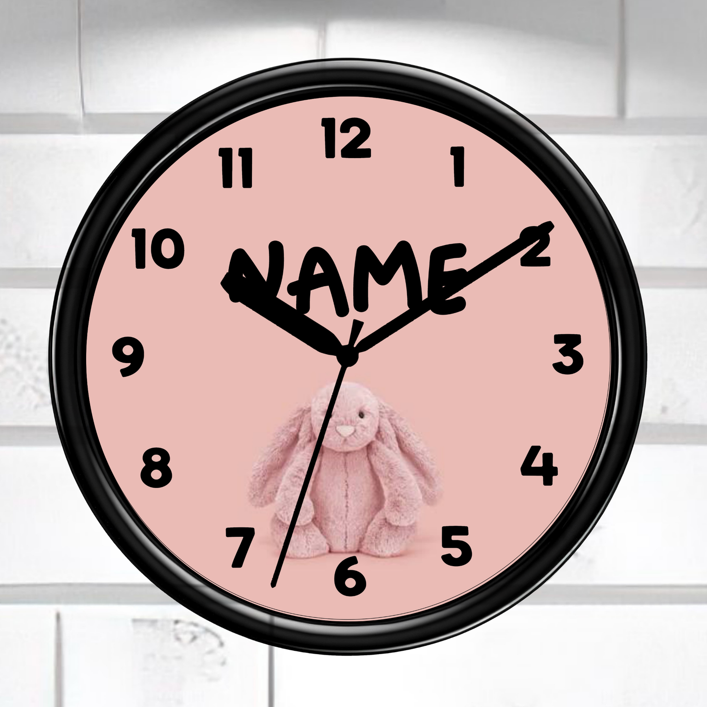 A round wall clock with a black frame and a pale pink face, featuring black numerals, black hands, a black script name placeholder, and a pink bunny illustration. The clock is mounted against a white tiled wall.