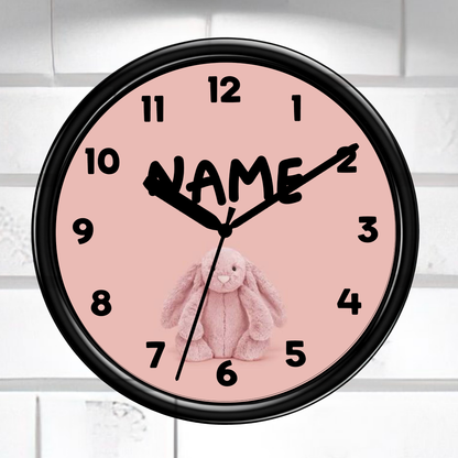 A round wall clock with a black frame and a pale pink face, featuring black numerals, black hands, a black script name placeholder, and a pink bunny illustration. The clock is mounted against a white tiled wall.