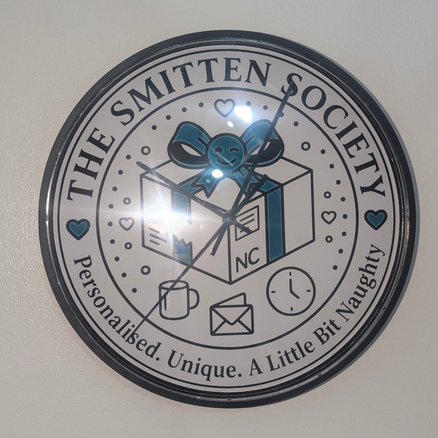 A round wall clock with a black frame and a white face featuring the text 'THE SMITTEN SOCIETY' and 'Personalised. Unique.