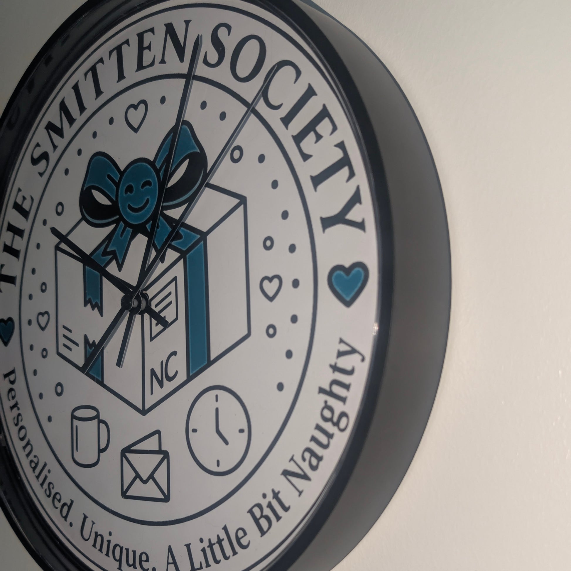 A round wall clock with a black frame and a white face featuring the text 'THE SMITTEN SOCIETY' and a central illustration of a gift box with a blue bow. The clock is mounted on a plain off-white wall.