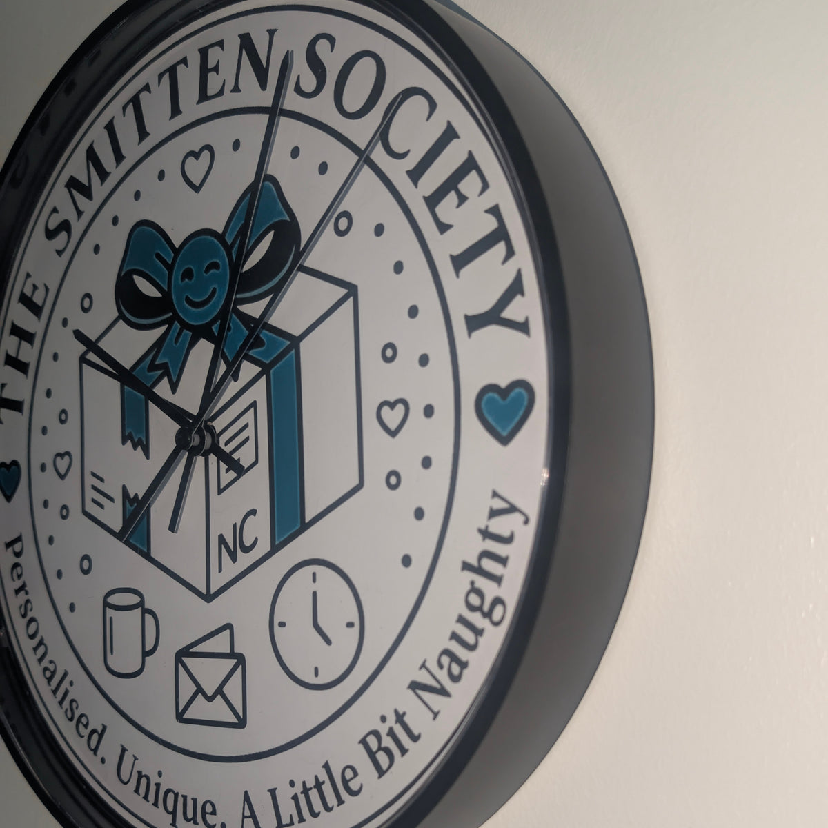 A round wall clock with a black frame and a white face featuring the text 'THE SMITTEN SOCIETY' and a central illustration of a gift box with a blue bow. The clock is mounted on a plain off-white wall.