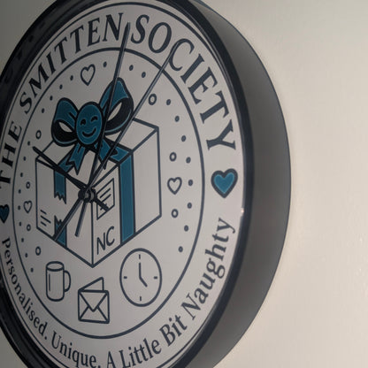 A round wall clock with a black frame and a white face featuring the text 'THE SMITTEN SOCIETY' and a central illustration of a gift box with a blue bow. The clock is mounted on a plain off-white wall.