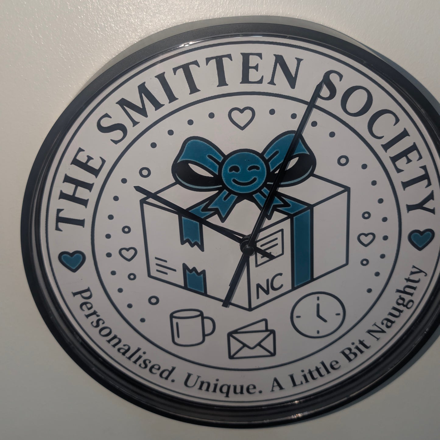 A round novelty wall clock with a black rim and a white face featuring a blue gift box with a smiley face ribbon, the text 'THE SMITTEN SOCIETY', and the phrase 'Personalised. Unique.