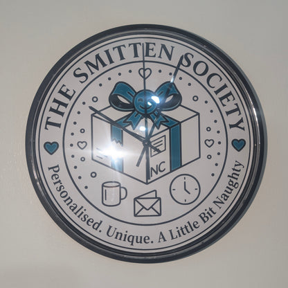 A round wall clock featuring a printed design of 'The Smitten Society' with a gift box, blue bow, coffee mug, envelope, and clock icons, mounted on a plain light wall.