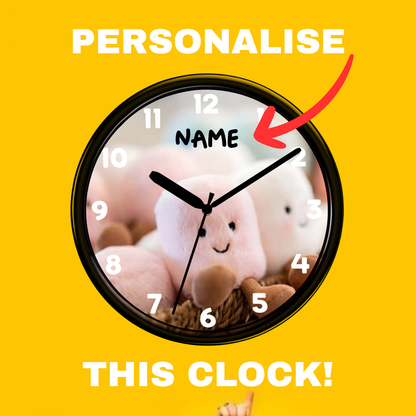 A customizable round wall clock with a black frame and a plush bunny photo-print face, shown on a bright yellow background with white text reading 'PERSONALISE THIS CLOCK!' and a red arrow pointing to the word 'NAME'.