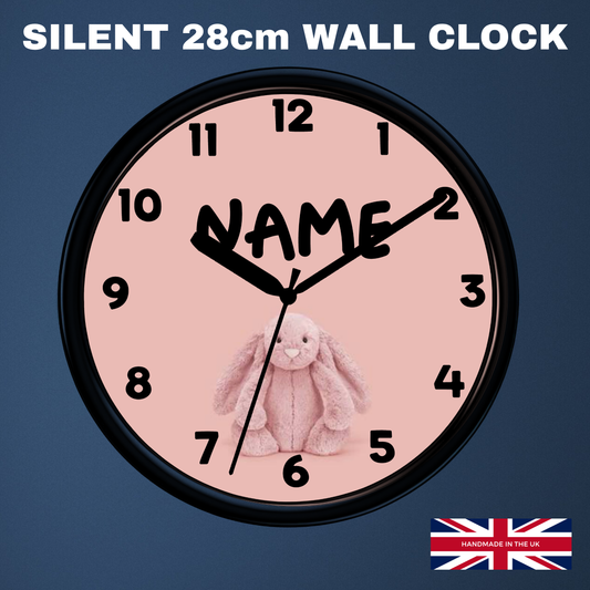 A round wall clock with a black frame and a pale pink face, shown against a dark blue background. The clock face has black numerals, black hands, and a customizable name printed across the center, with a small pink bunny illustration near the bottom.