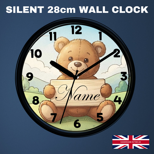 A round wall clock with a black frame featuring a teddy bear illustration holding a sign that says 'Name'. The clock is shown against a dark blue background with the text 'SILENT 28cm WALL CLOCK' at the top and a small 'Handmade in UK' flag graphic in the bottom right corner.