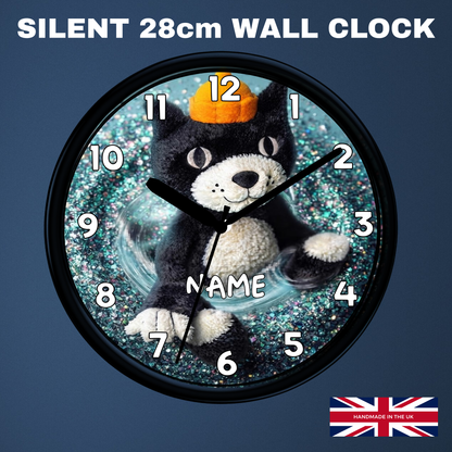 A round wall clock with a black cat plush figure in the center, wearing an orange pumpkin hat, on a glittery blue background. The clock has a black frame and white Arabic numerals.