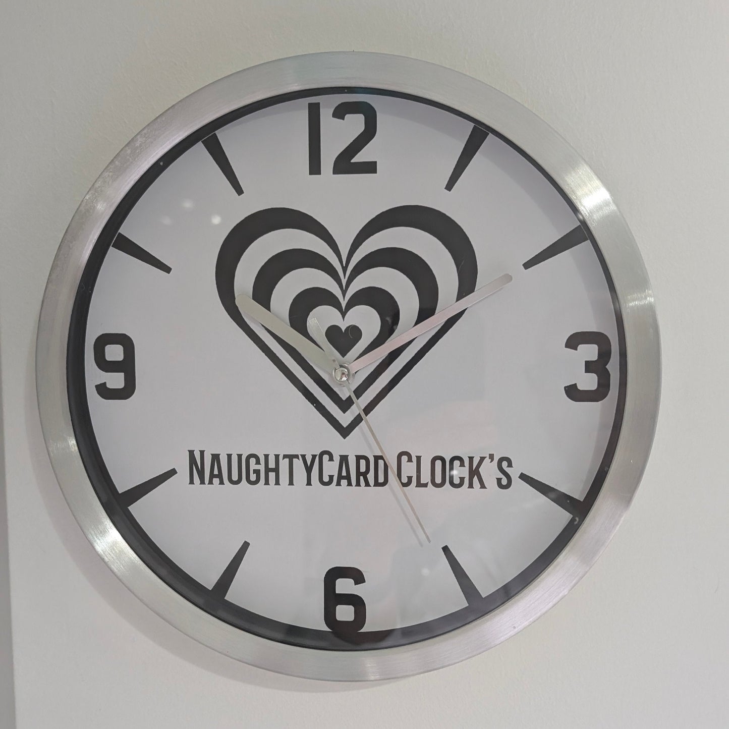 A silver metallic wall clock featuring an image of NaughtyCard logo and numbers, predominantly in black and white with a silver or metallic accent. Metallic Wall Clock from NaughtyCard