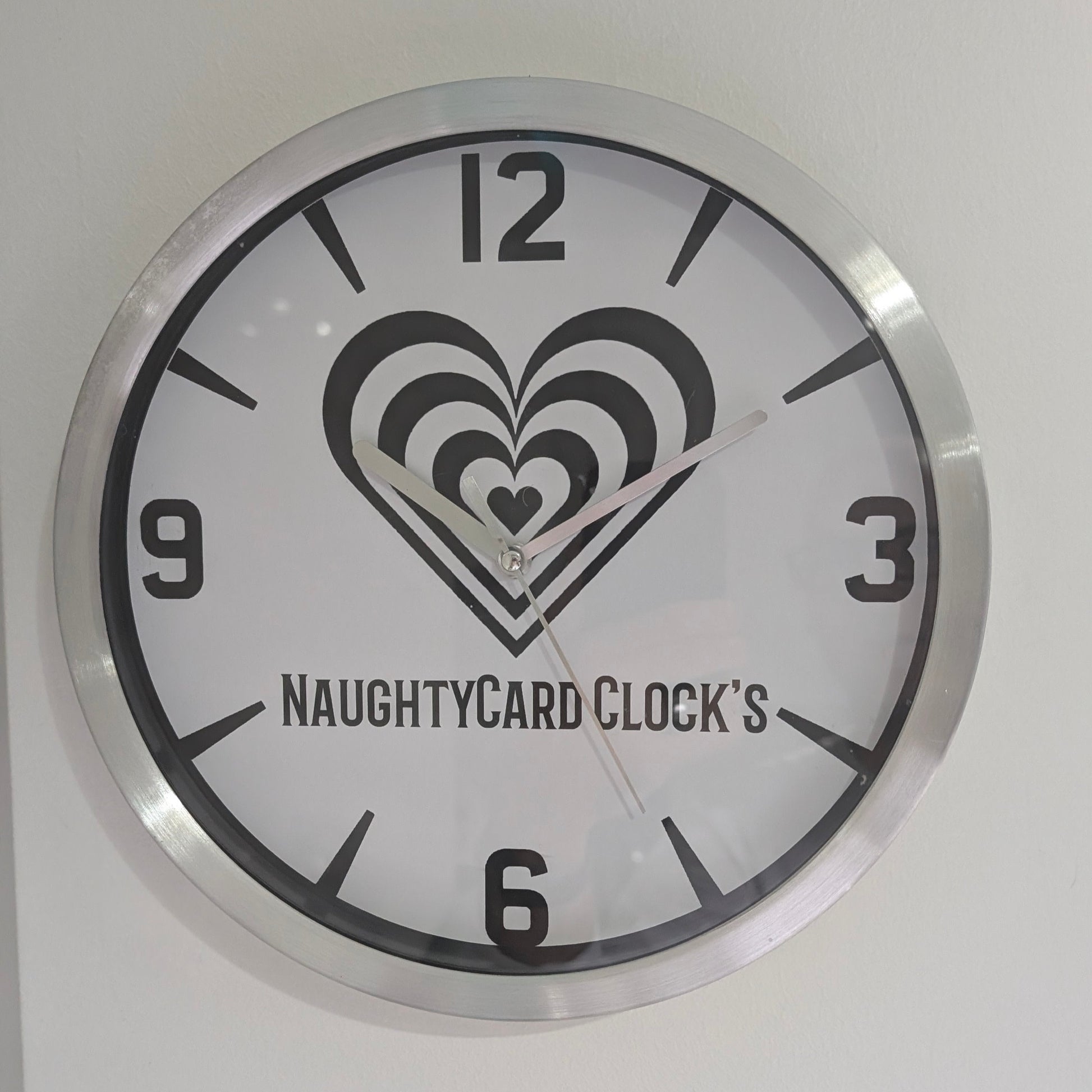 A silver metallic wall clock featuring an image of NaughtyCard logo and numbers, predominantly in black and white with a silver or metallic accent. Metallic Wall Clock from NaughtyCard